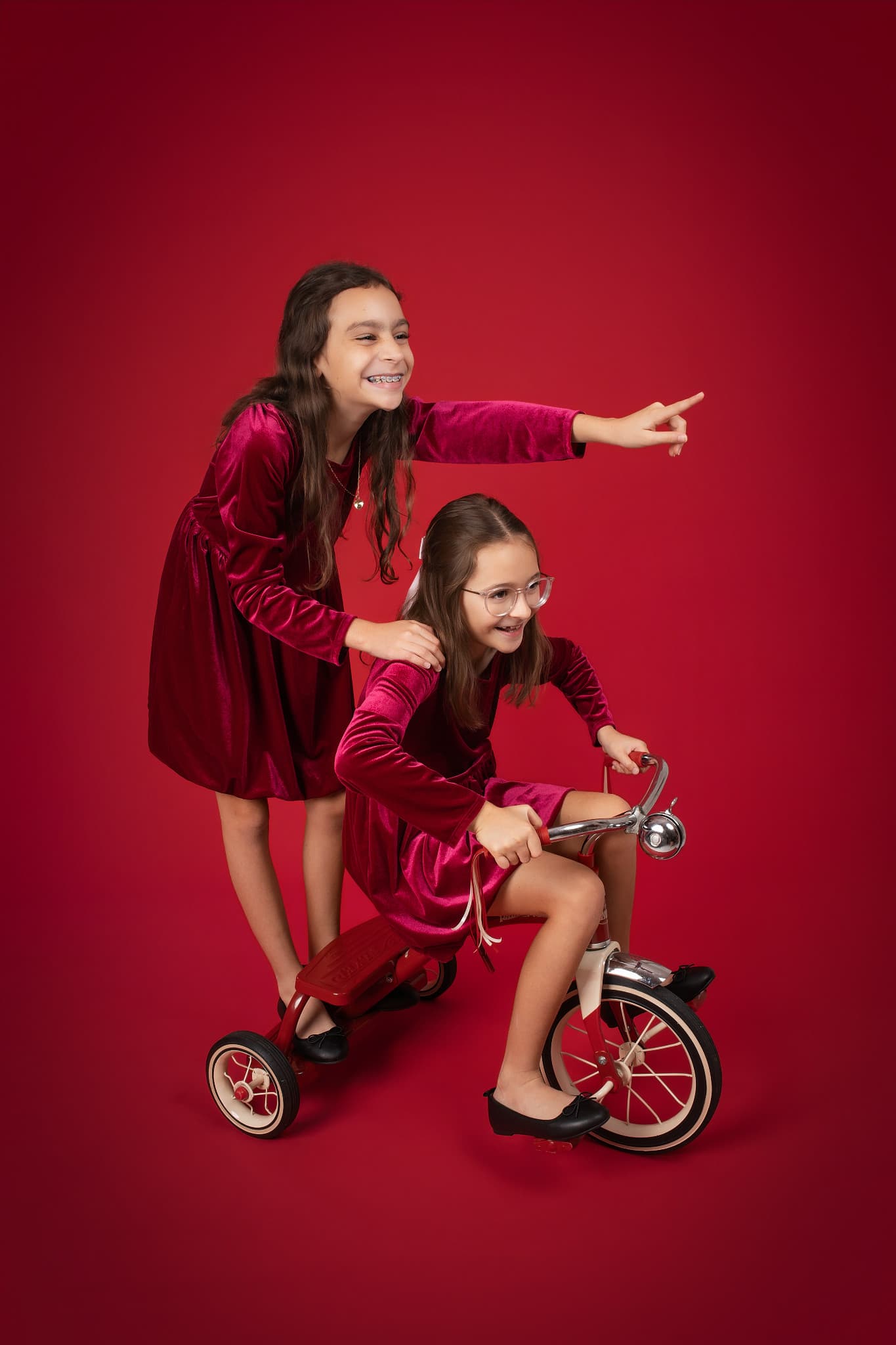 Two sisters in red velvet dresses with a vintage tricycle against a deep red backdrop