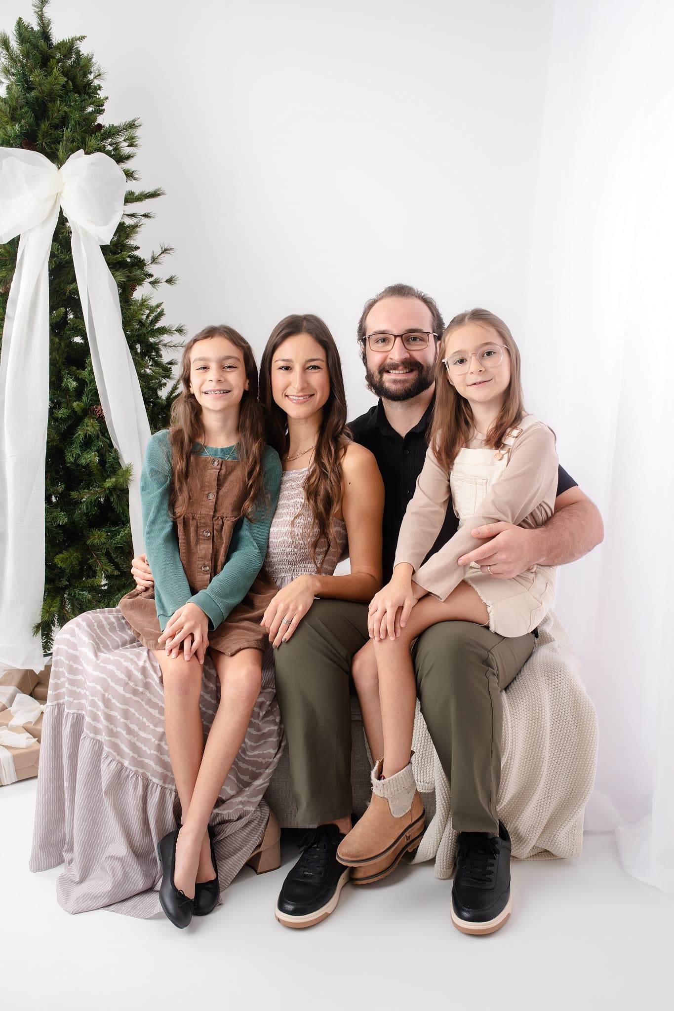 A family of four on a bench beside a white Christmas tree tied with a wide white bow